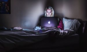 Person with shoulder length blonde hair sitting in a dark room in a bed, looking at their laptop. Photo by Photo by Victoria Heath on Unsplash