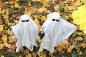 two people in white sheets and sunglasses, dressed as ghosts, shot from above as they stand on autumn leaves