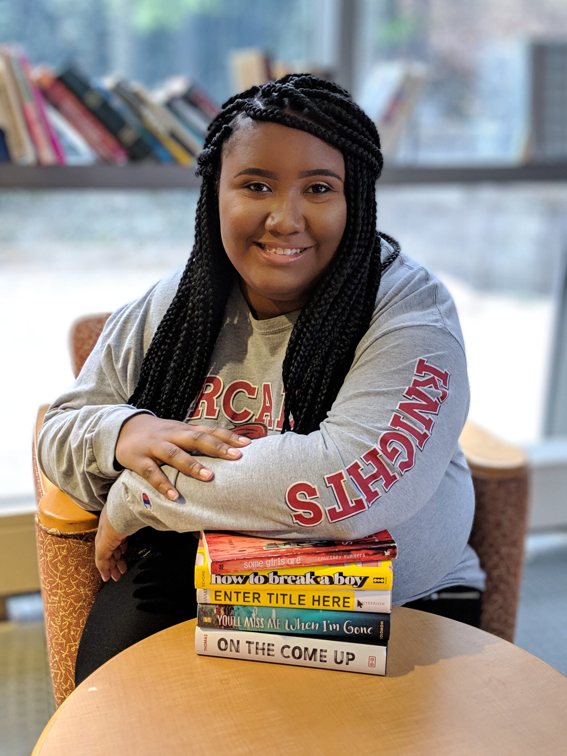 headshot of Sierra Elmore, a Black woman with long hair, smiling and sitting at a table next to a pile of books