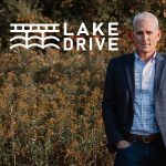 Photo of a white man with gray hair in a button up and suit jacket standing in a field. The logo for Lake Drive is set next to his head, which includes text beside a line drawing of a bridge over a lake.