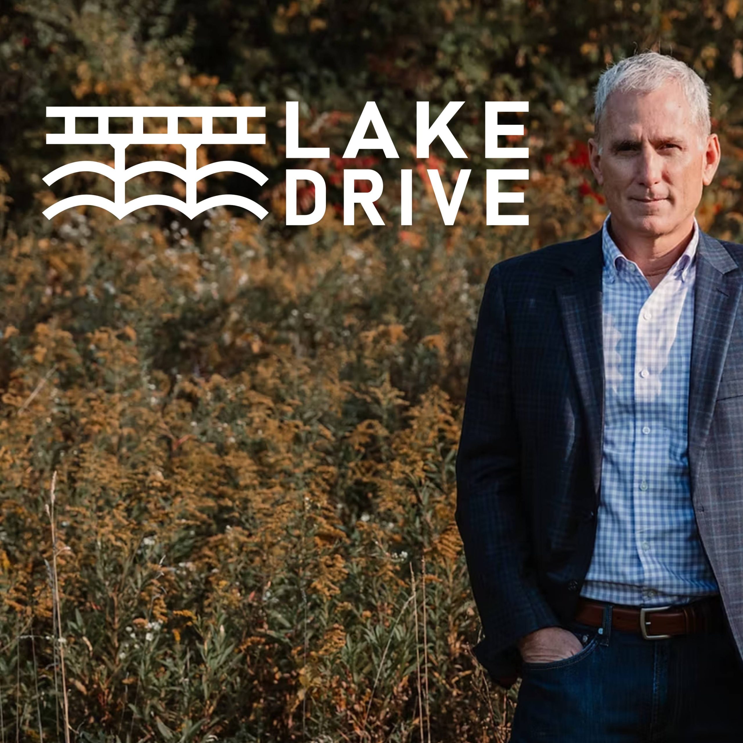 Photo of a white man with gray hair in a button up and suit jacket standing in a field. The logo for Lake Drive is set next to his head, which includes text beside a line drawing of a bridge over a lake.